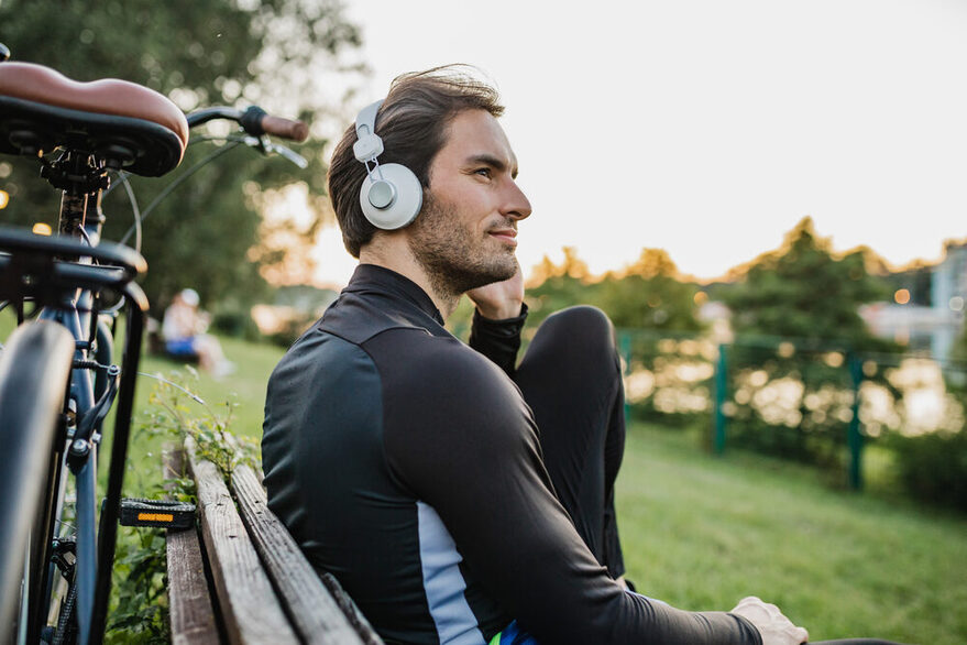 Junge Sportler macht auf einer Bank eine Pause vom Radfahren am See und hat Kopfhörer auf