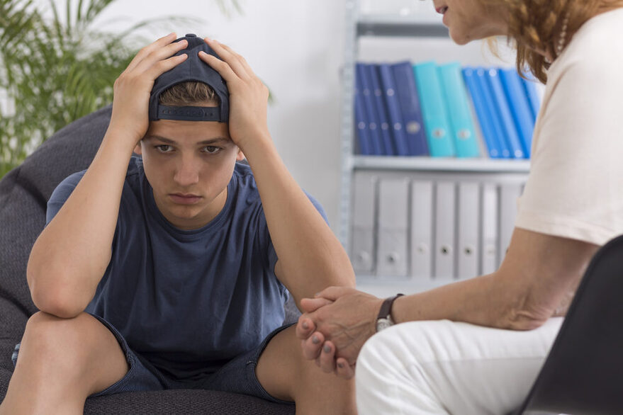 Ein Junge sitzt nachdenklich in einem Büro und hält die Hände über dem Kopf. Ein Frau redet mit ihm.