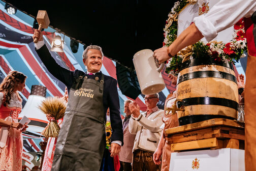 ein Mann mit lederner Schürze recket einen Holzhammer in die Luft. Neben ihm steht ein Bierfass.