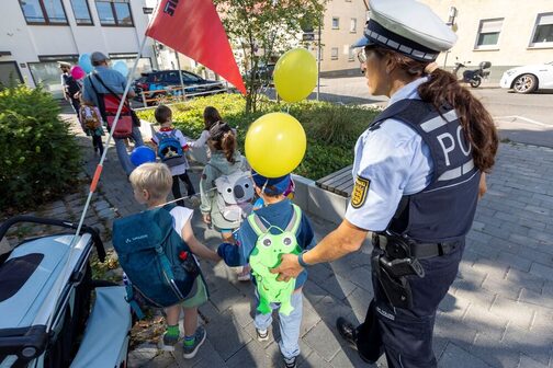 Eine Polizeibeamtin führ einige Schulkinder über einen Bürgersteig. Die Polizistin trägt Uniform, die Kinder jeweils einen Rucksack auf dem Rücken.