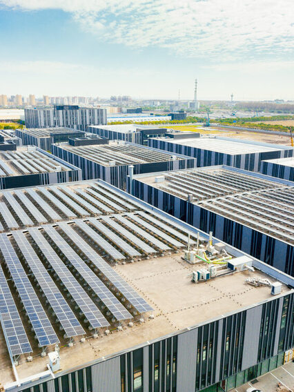 Im Vordergrund sind moderne Gebäude mit Solaranlagen auf dem Dach zu sehen. Im Hintergrund ist eine Stadt zu erkennen.