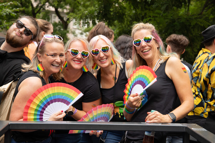 Vier Frauen mit Regenbogen-Fächern und Herz-Sonnenbrillen lachen in die Fotokamera.