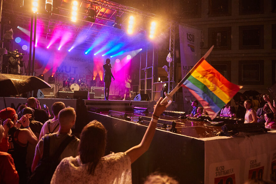 Musik-Act auf Bühne, davor schwenkt jemand eine Regenbogen-Fahne.