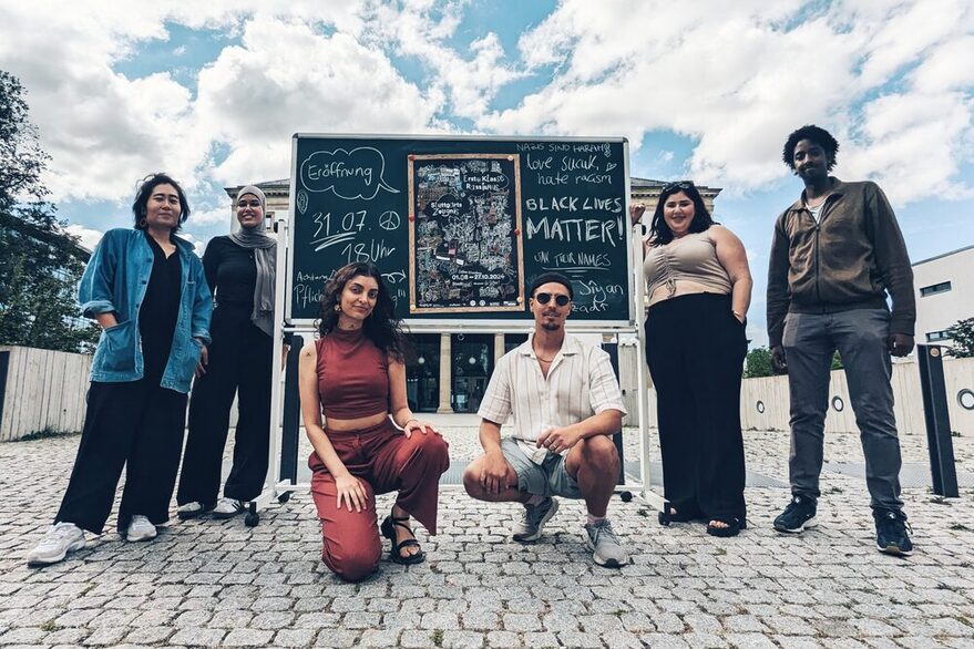 Gruppenfoto vor einer tAfel m it dem Plakat zur Ausstellung "Erste Klasse Rassismus" im StdtPalais Stuttgart.