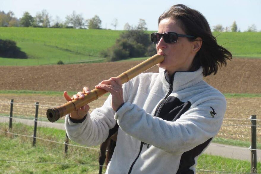 Sylvia Shoin-Bittmann (Sonnenbrille, dunkle mittellange Haare, weiße Fleecejacke) spielt im Freien Shakuhachi, im Hintergrund hügeliges Ackerland