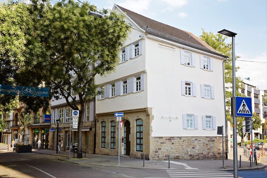 Eine Fotografie zeigt die Fassade und den Eingang des Hegel-Hauses an der Eberhardstraße an einem sonnigen Sommertag. Links im Bild ist ein grüner Baum entlang der Eberhardstraße zu sehen, mit Blickrichtung vom Tagblattturm.