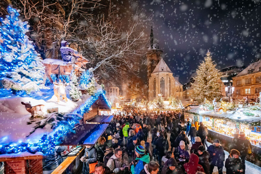 Draufsicht im Dunkeln auf einen geschmückten, beleuchteten Weihnachtsmarkt und Gebäuden mit Passanten bei Schneetreiben.